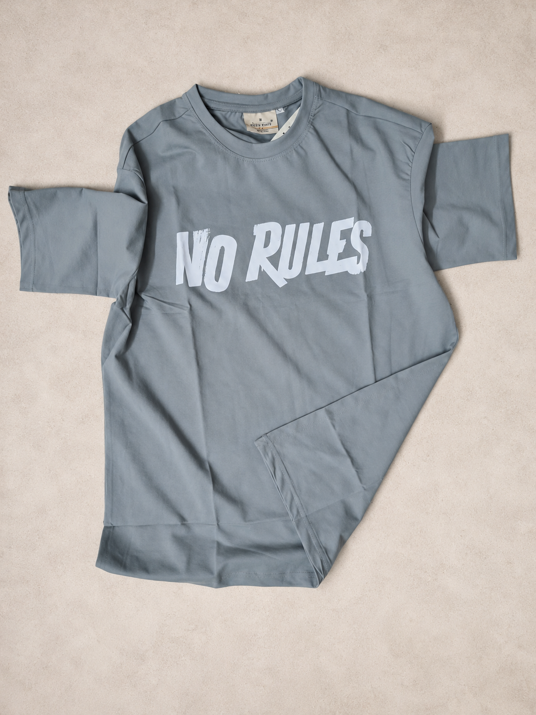 A slate gray long-sleeve bodysuit with the text 'NO RULES' printed in white across the chest, laid flat on a textured beige background.